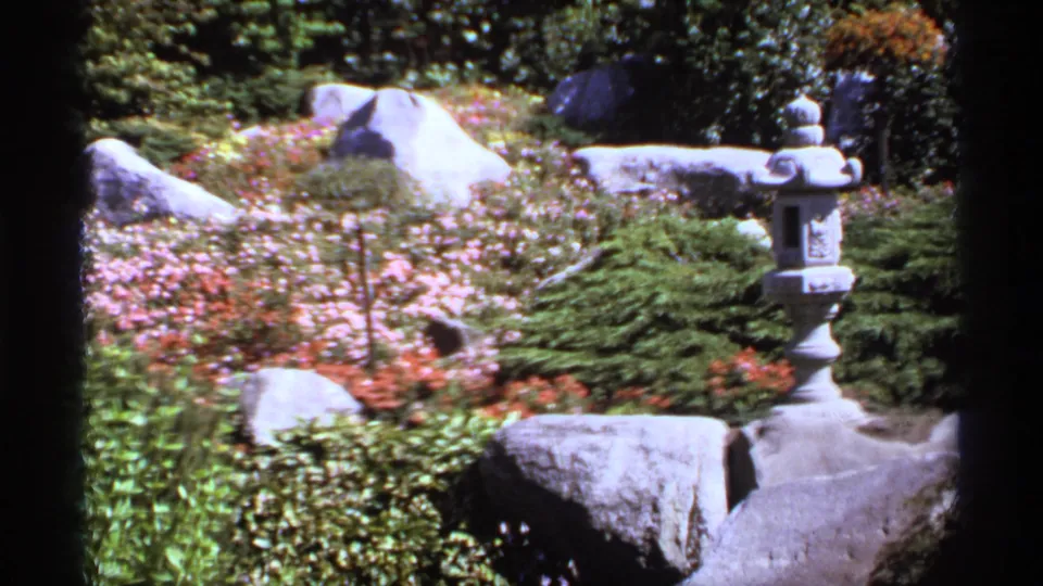 1974 Canada 8mm frame showing a Japanese-style garden with a stone lantern, boulders, shrubs, and pink flower beds.