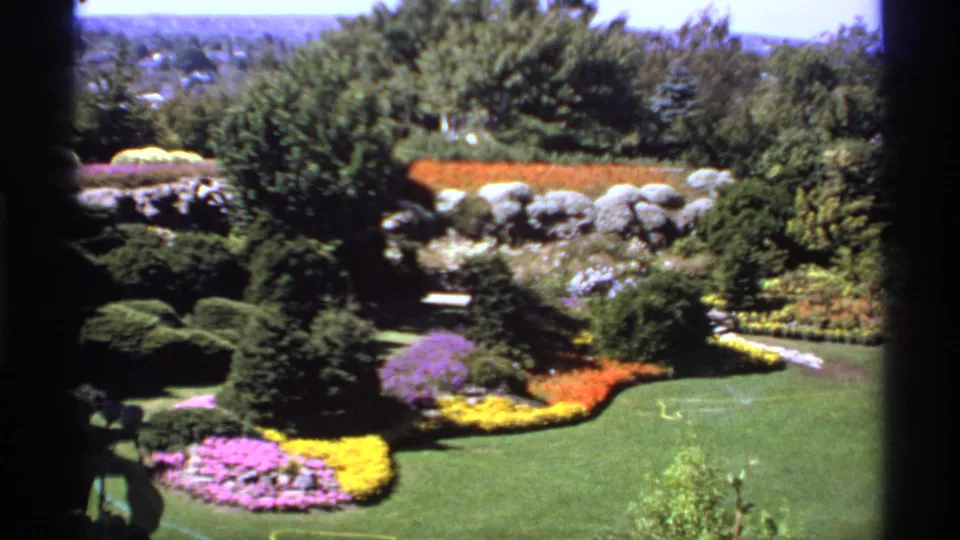 1974 Canadian 8mm wide view of a landscaped garden with patterned yellow, orange, and purple flower beds around a green lawn.