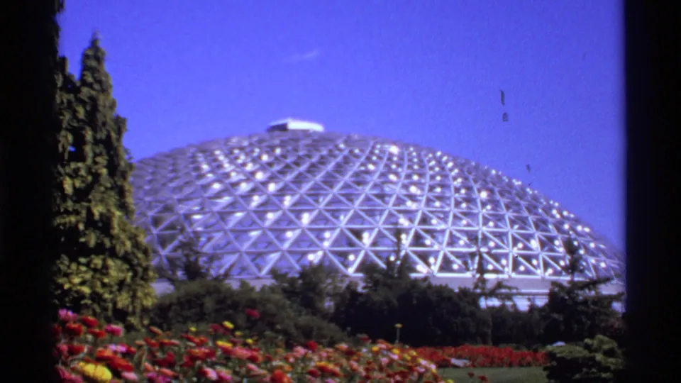 1974 Canada frame showing a geodesic dome behind flower beds, evergreens, and a deep blue sky in a landscaped garden.
