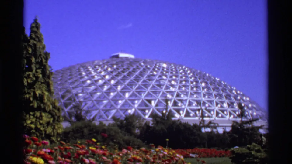1974 Canada home-movie frame of a geodesic dome and landscaped flower beds, with trees at the edges and saturated blue sky above.