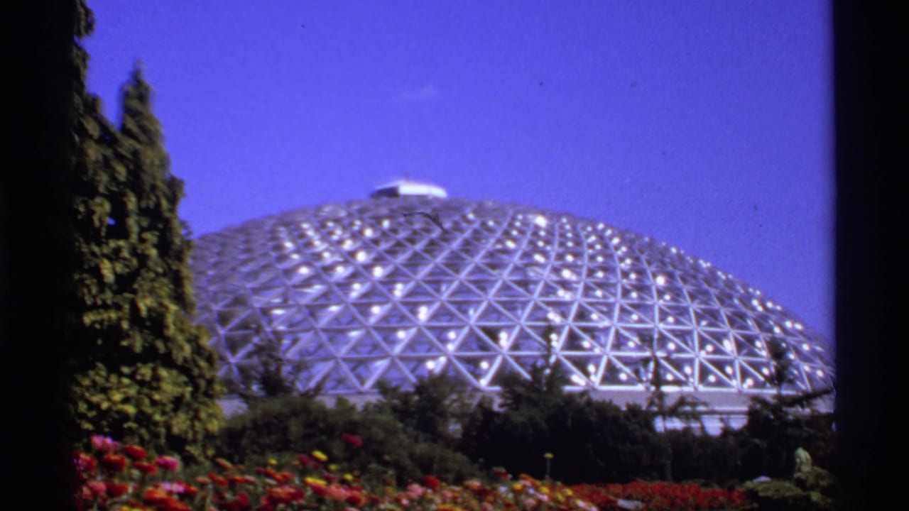 Geodesic glass dome behind flowerbeds and evergreens under a blue sky.