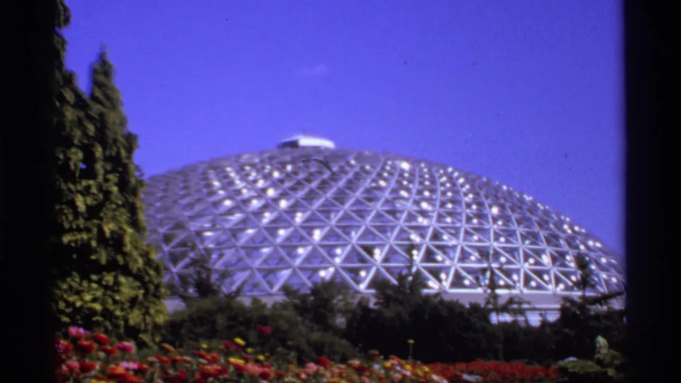 1974 Canadian 8mm frame showing a geodesic glass dome in a garden, framed by evergreens and colorful flowerbeds.