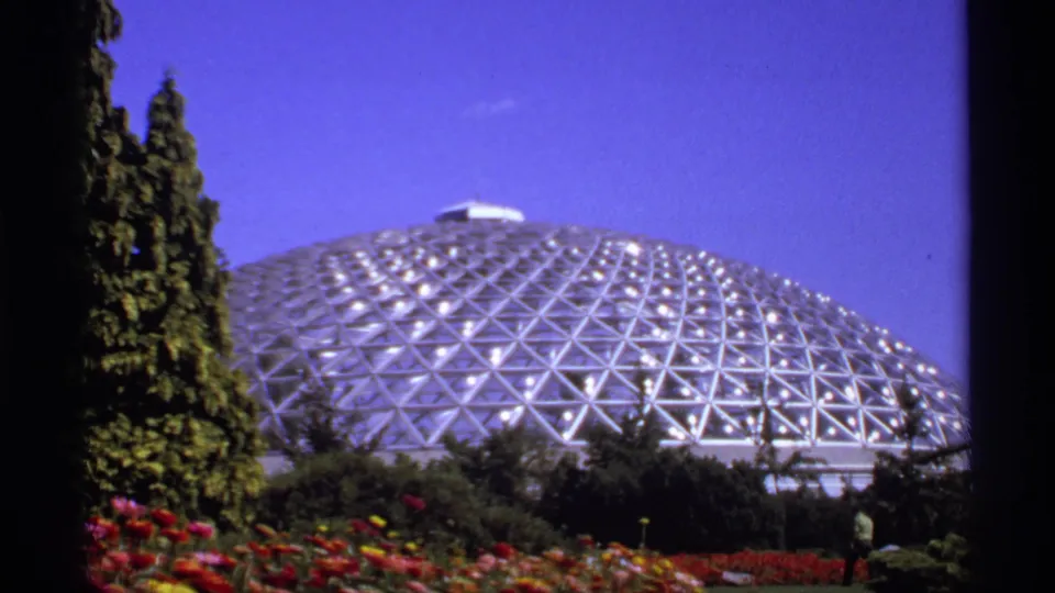 1974 Canadian 8mm view of a geodesic greenhouse-like dome rising behind trees, flowers, and a small distant visitor.