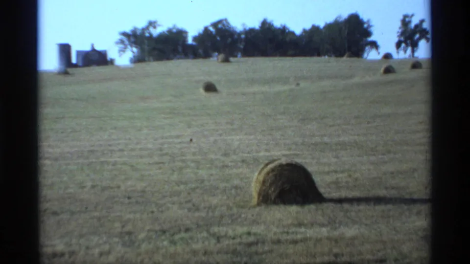 Wide 1974 Minnesota farm landscape with scattered hay bales, distant trees, and farmhouse structures along the horizon.