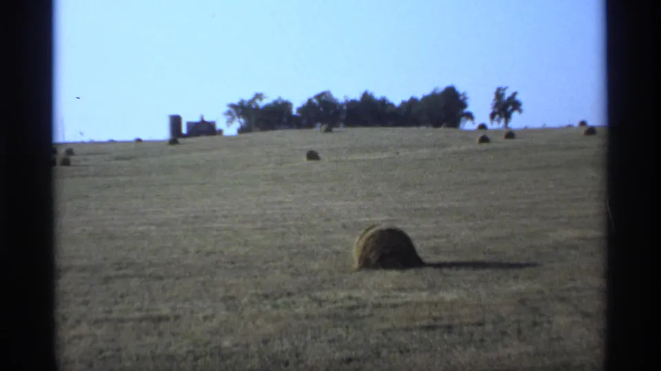 1974 Minnesota 8mm frame of open rolling farmland with a large round hay bale in front and distant farm buildings near a tree line.