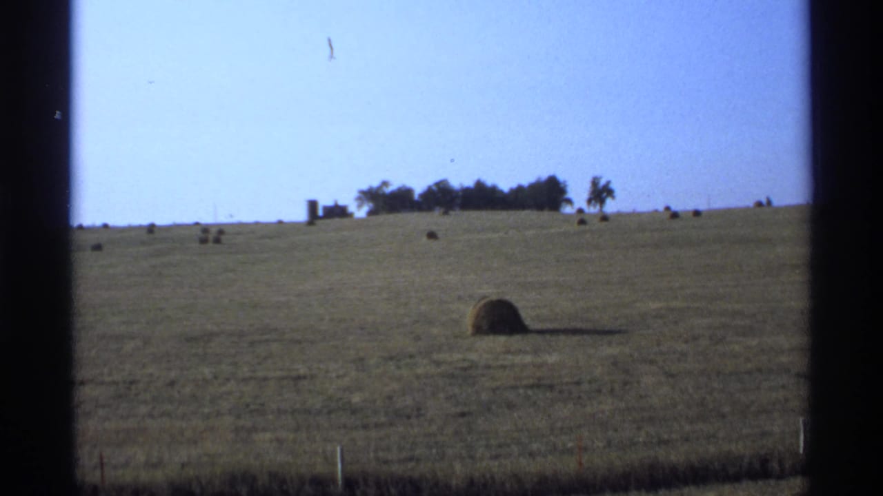 Open farmland with hay bales, a distant building cluster, trees, and blue sky.