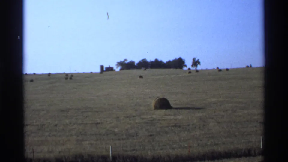 1974 Minnesota rural landscape frame showing hay bales scattered across a harvested field below distant trees and farm structures.