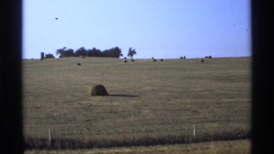 1974 Minnesota 8mm still of a broad rural field with round hay bales, distant trees, and farm structures near the left horizon.