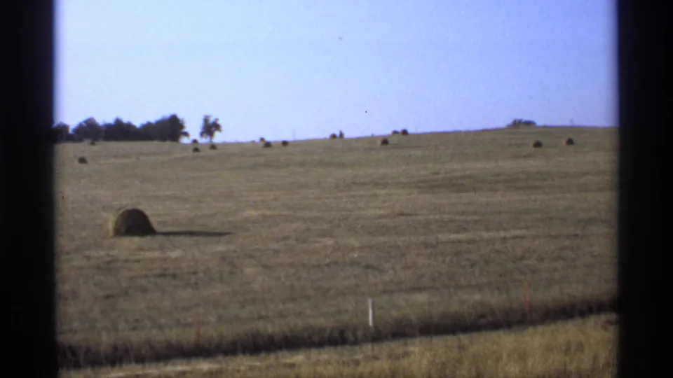 1974 Minnesota frame of rolling farmland with a round hay bale in the foreground, fence posts below, and trees on the far ridge.