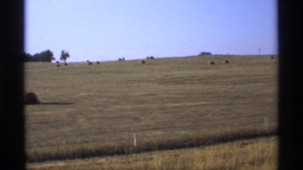 1974 Minnesota rural field with distant hay bales, low fence posts, roadside grass, and dark window-frame edges.