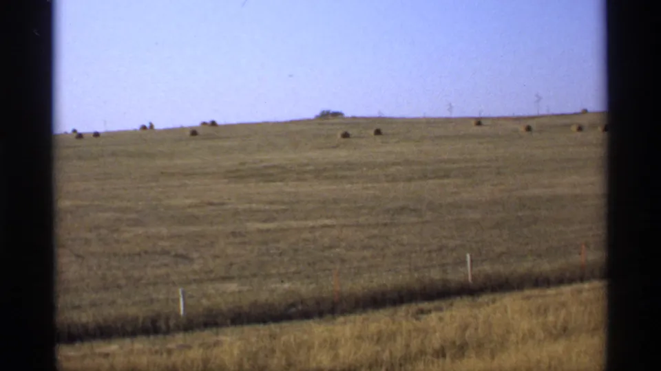 1974 Minnesota farmland viewed from a moving vehicle, with hay bales across the horizon and a simple fence line in front.