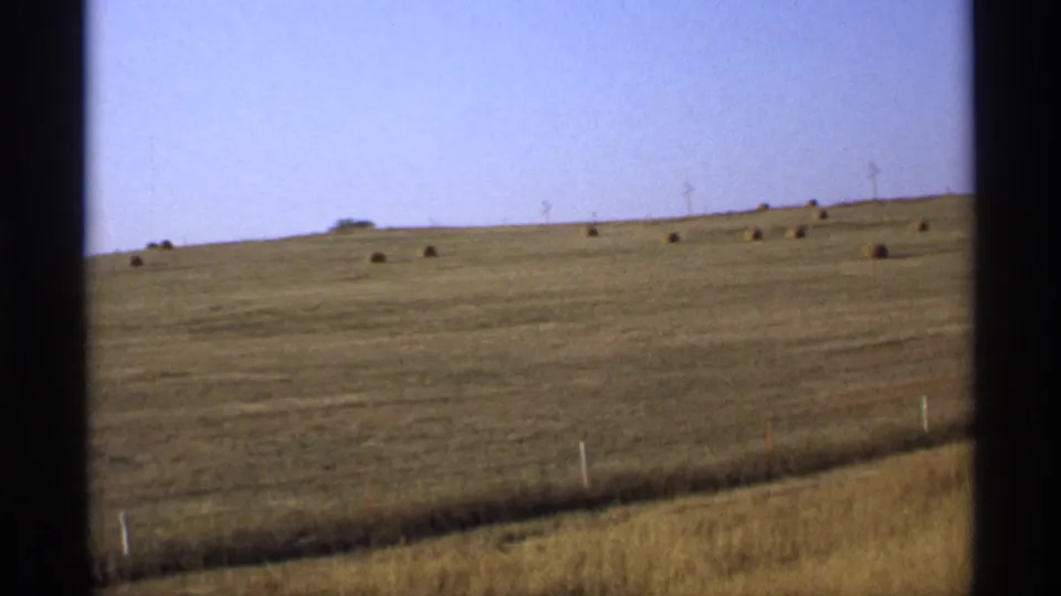 1974 Minnesota field with a sloped horizon, scattered hay bales, roadside fence posts, and visible 8mm film grain.