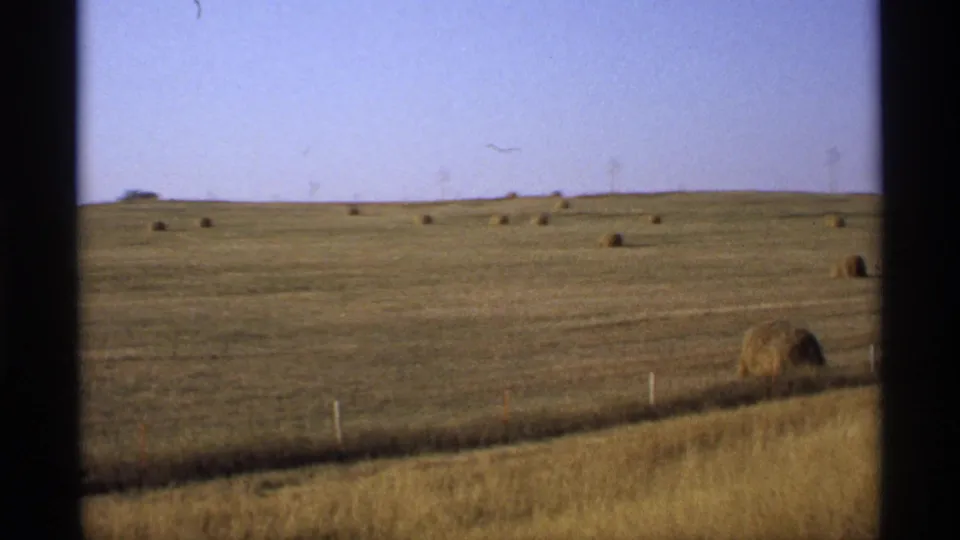 1974 Minnesota end-of-summer field with golden foreground grass, scattered hay bales, and dark window-frame borders.