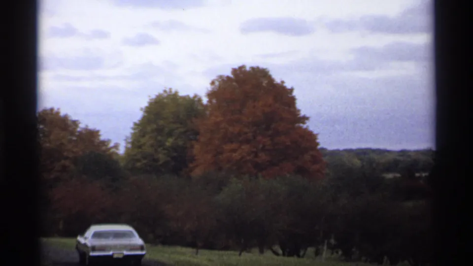 Minnesota 1974 home-movie view of a car near a rural road, with orange and green fall trees and a cloudy sky.