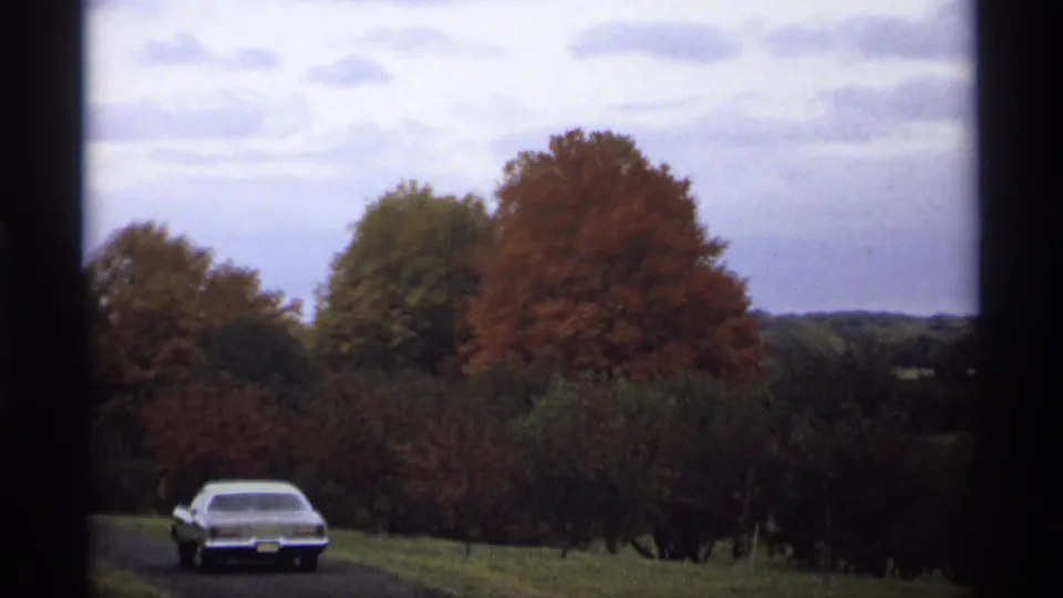 1974 Minnesota 8mm frame showing a light-colored sedan on a rural road with red and green autumn trees beyond a dark window edge.