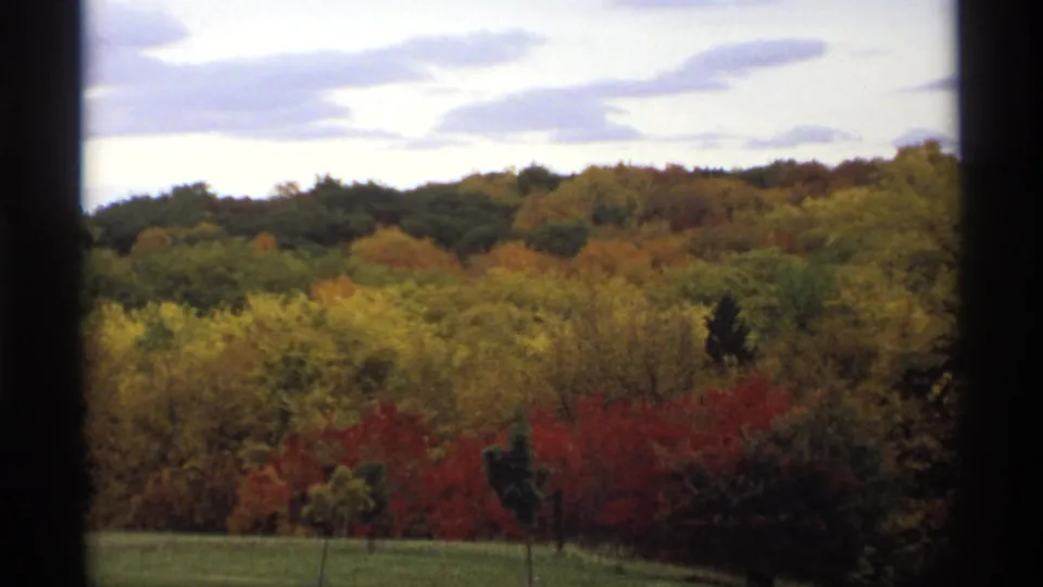 1974 Minnesota 8mm frame showing a distant autumn hillside with mixed red, yellow, and green foliage below a cloudy sky.