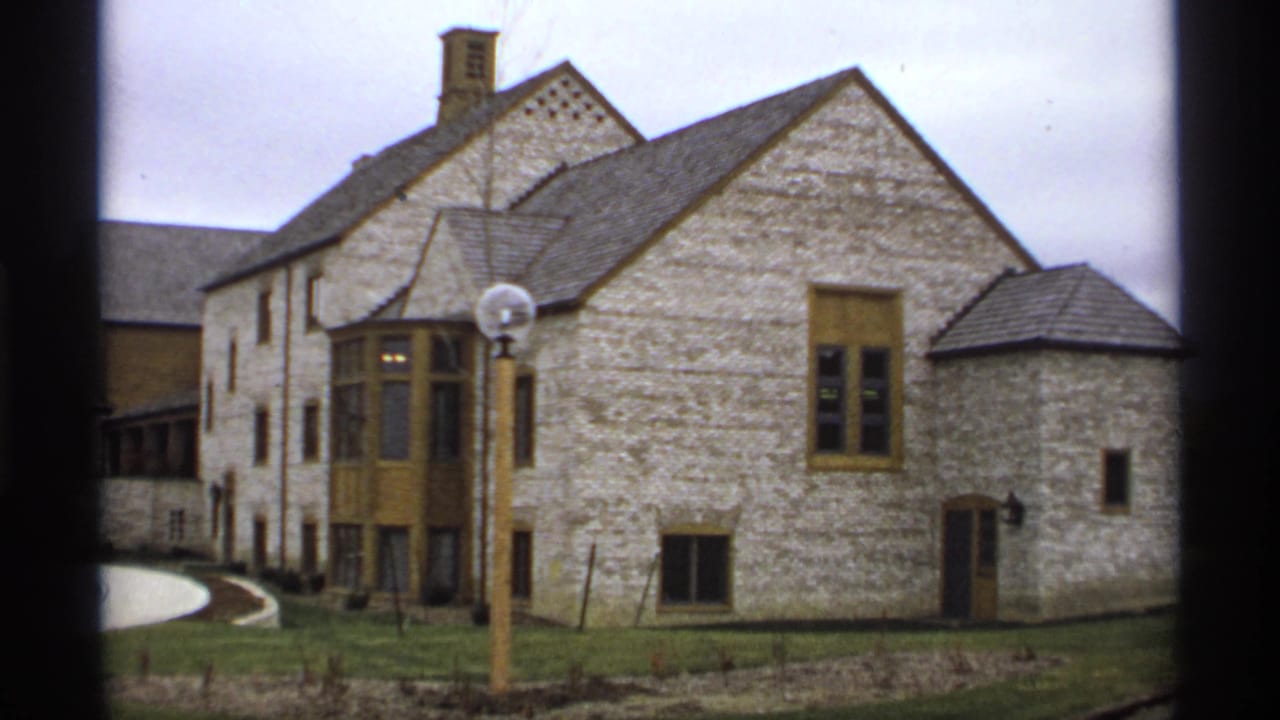 1974 Minnesota home-movie frame holding on the stone building facade, tan trim, foreground globe lamppost, and grounds.