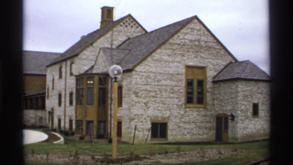 1974 Minnesota home-movie frame holding on the stone building facade, tan trim, foreground globe lamppost, and grounds.