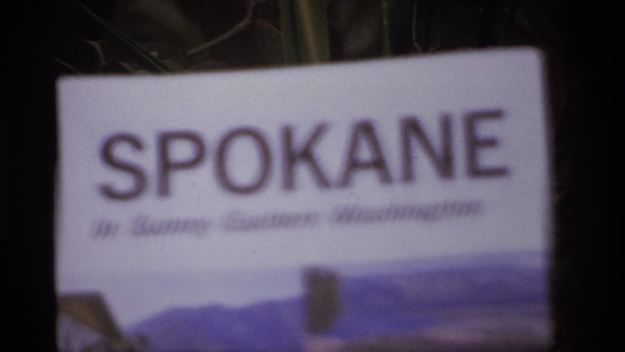 1974 8mm frame showing a soft-focus Spokane pamphlet held or placed outdoors with dark trees in the background.