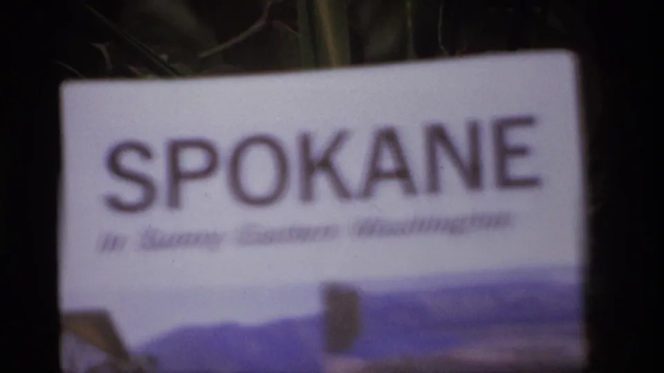 1974 8mm frame showing a soft-focus Spokane pamphlet held or placed outdoors with dark trees in the background.