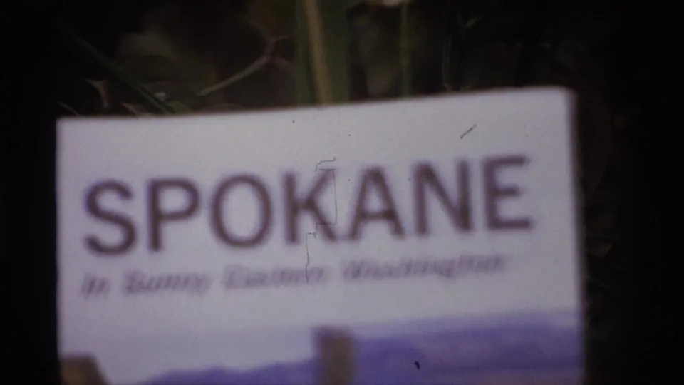 1974 8mm frame with the word Spokane on a travel pamphlet, partially soft and backed by dark outdoor foliage.