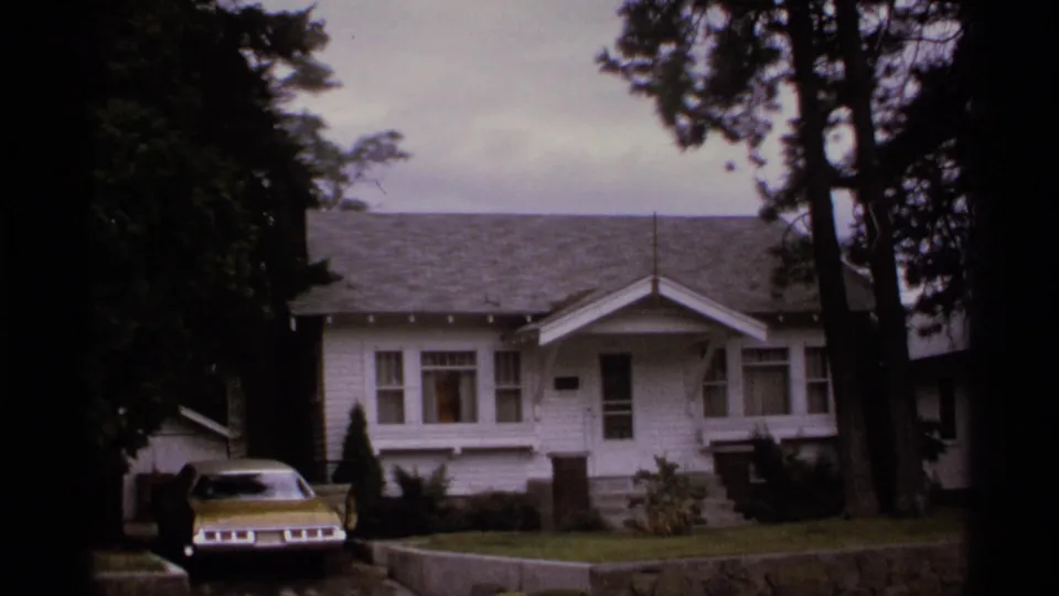 1974 Spokane home-movie frame showing a white house, front lawn, tall trees, and a yellow car parked near the driveway.