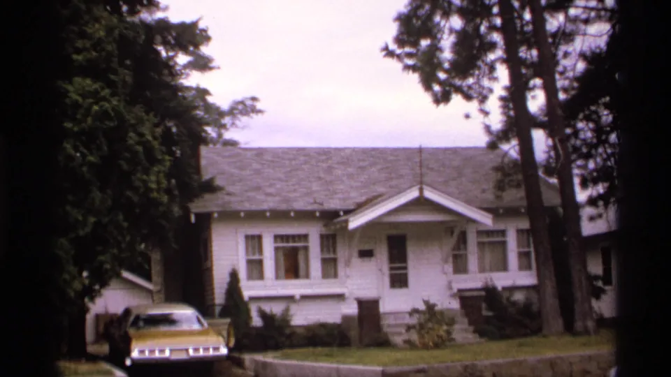 1974 Spokane residential exterior with a white house, yellow car, lawn, driveway, and tall trees in soft 8mm focus.