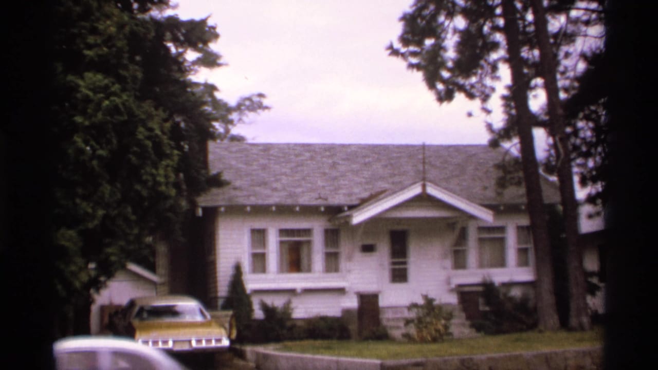 White bungalow with a gold car parked in the driveway among tall trees.