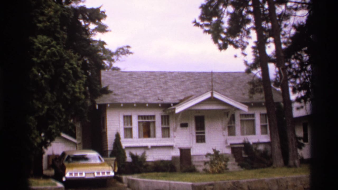 1974 Spokane residential exterior with a white bungalow, gold sedan, divided front windows, lawn, and tall trees partly framing the view.