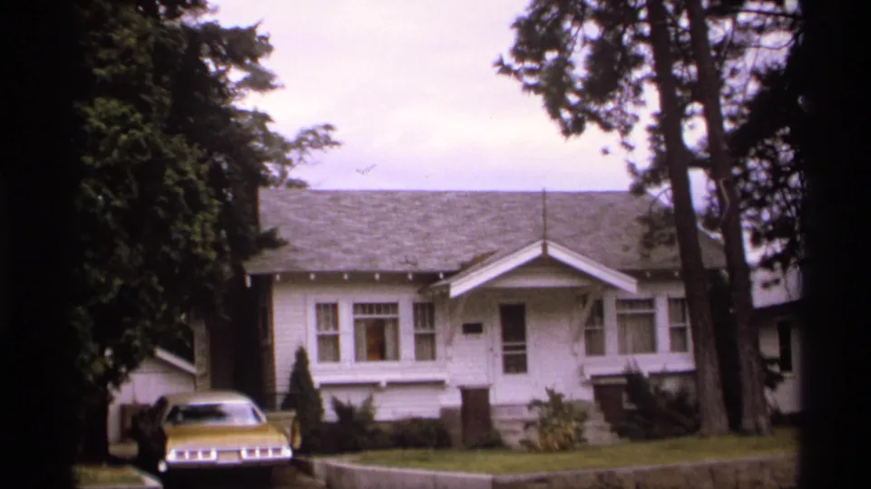 1974 Spokane frame of a white single-story bungalow set behind a low stone wall, with a gold car in the driveway and tall trees around the property.