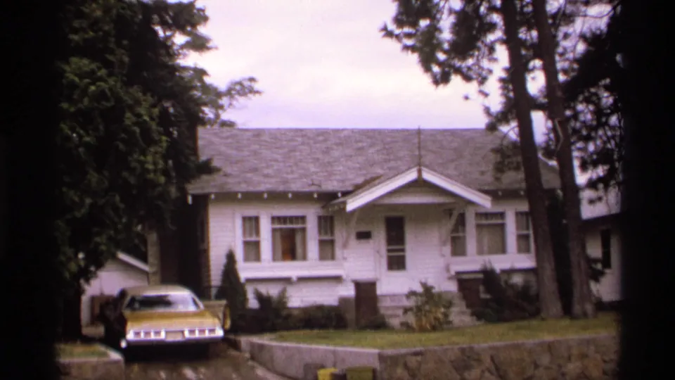 1974 Spokane home exterior framed by dark tree trunks, showing a white bungalow, front porch, lawn, low wall, and gold car parked near the driveway.