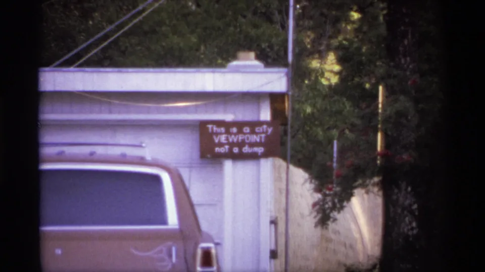 Spokane 1974 home-movie frame with a brown car in front of a white garage, leafy trees, a fence, and the city viewpoint sign.