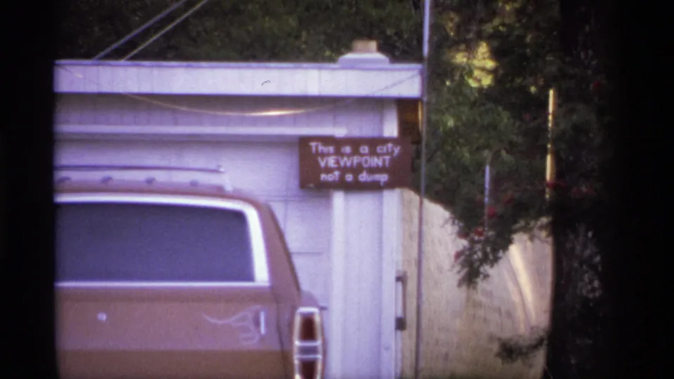 1974 Spokane frame showing the rear of a brown car, white garage door, trees, fence, and the sign marking a city viewpoint rather than a dump.
