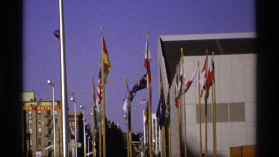 Visitor-shot 8mm view of Expo '74 flags, flagpoles, streetlights, and a modern building in Spokane.