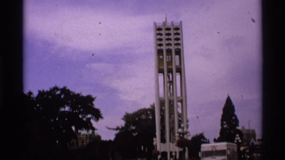 1974 Vancouver home-movie frame with the bell tower centered above trees, soft grain, and a partial white van at street level.
