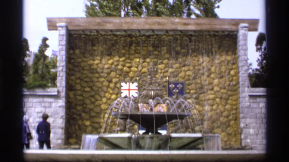 1974 Vancouver home-movie frame of a public fountain with cascading water, a rounded-stone backdrop, flags, and visitors walking nearby.