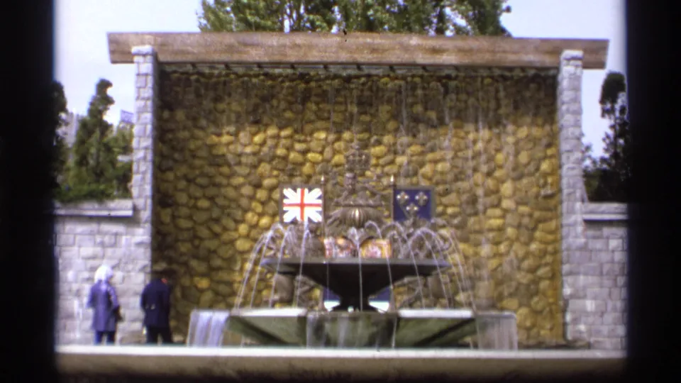 1974 Vancouver frame showing a fountain with arcing water, stone wall backdrop, Union Jack and blue flag details, and visitors nearby.