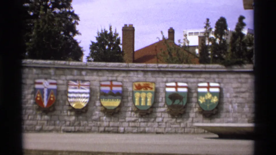 1974 Vancouver home-movie frame showing six colorful Canadian shields on a gray stone wall.
