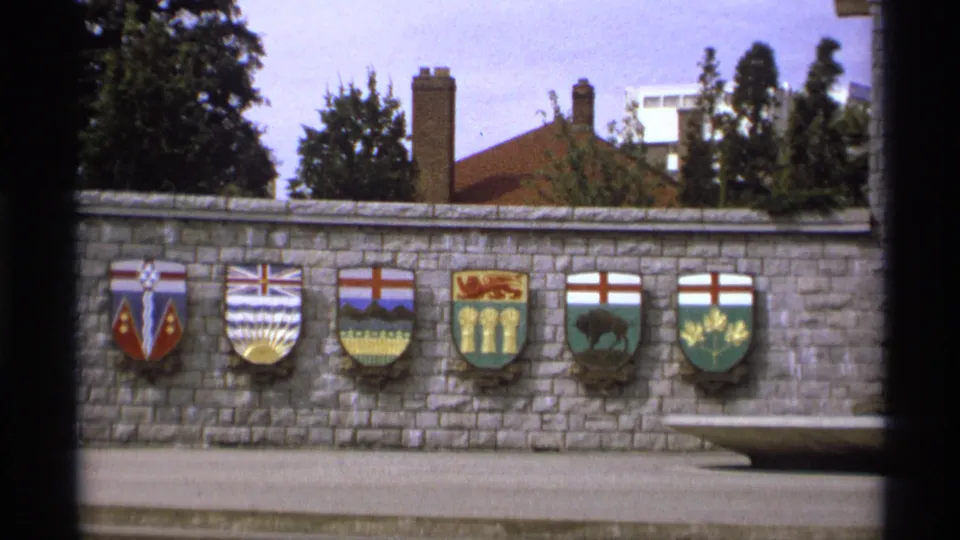 1974 Vancouver frame holding on a wall of colorful shields with trees and rooftops in the background.