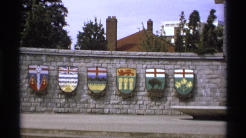1974 Vancouver 8mm frame showing a grey stone wall lined with colorful Canadian coats of arms, seen through dark contact-sheet borders.