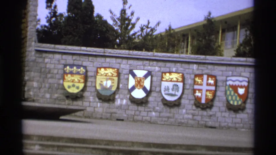 1974 Vancouver 8mm frame focusing on several coats of arms mounted along a stone wall, with trees and a building behind it.