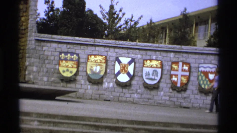 1974 Vancouver 8mm frame showing heraldic shields on a stone wall, steps in the foreground, and a partial pedestrian at the right edge.
