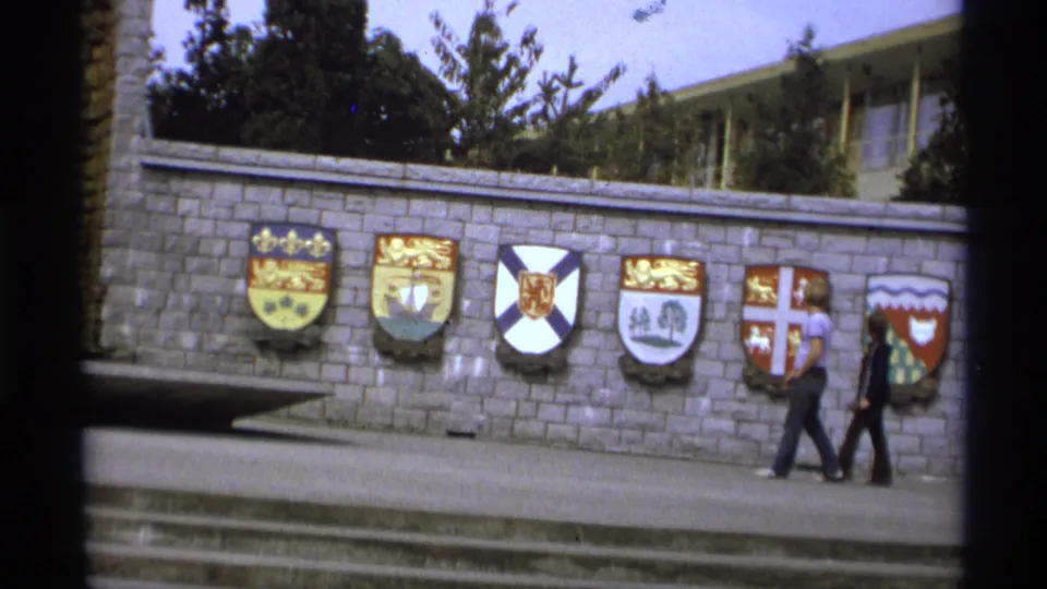 1974 Vancouver 8mm frame with two pedestrians passing a grey stone wall lined with colorful coats of arms and steps in the foreground.