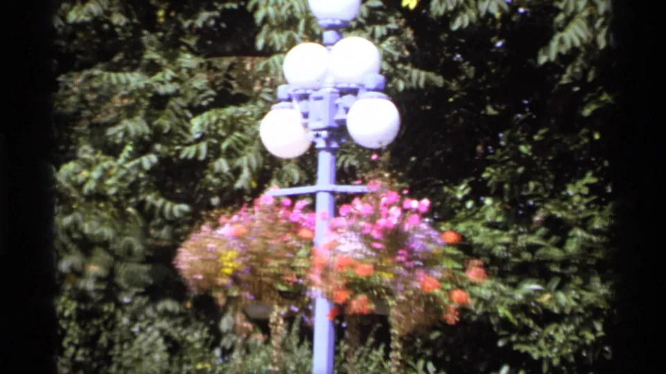 Final 1974 Vancouver frame showing a decorative lamppost with globe lights and hanging flowers against dense trees.