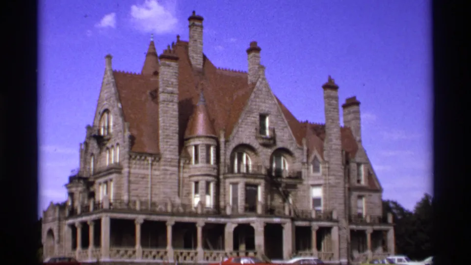 1974 Vancouver 8mm frame showing an ornate stone building with red-brown roof, multiple chimneys, arched windows, porch, and parked cars.