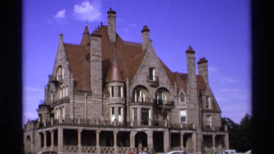 1974 Vancouver 8mm frame with a closer angled view of the stone building, showing gables, chimneys, arched windows, porch railing, and cars at the base.
