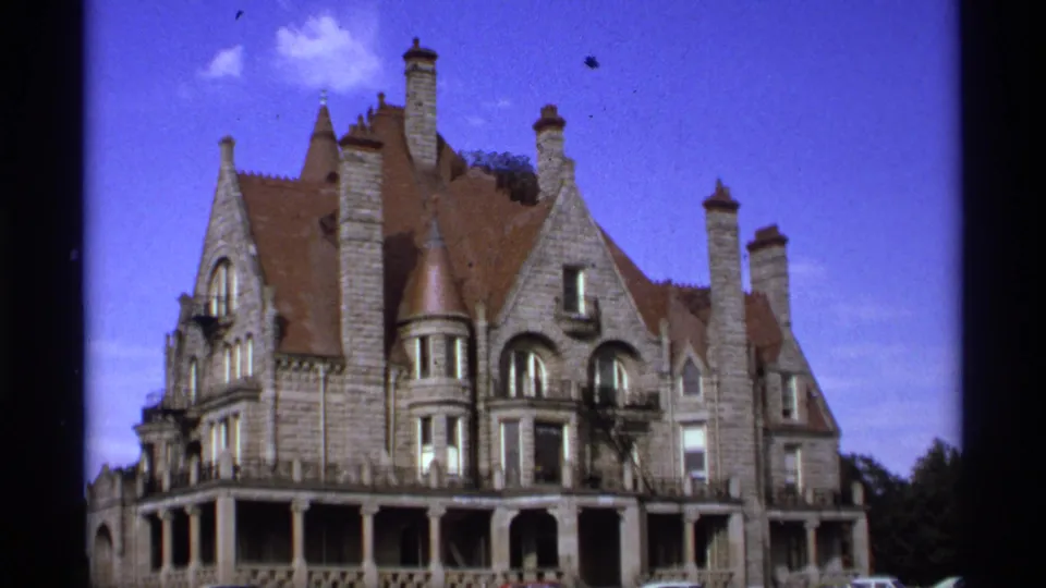 1974 Vancouver 8mm frame showing the ornate stone building from a steady exterior view, with chimneys, porch arches, arched windows, blue sky, and slight film marks.