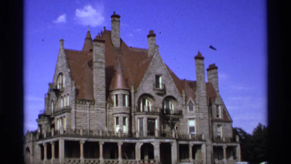 1974 Vancouver home-movie frame showing a large grey stone building with steep red-brown rooflines, chimneys, and arched upper windows.