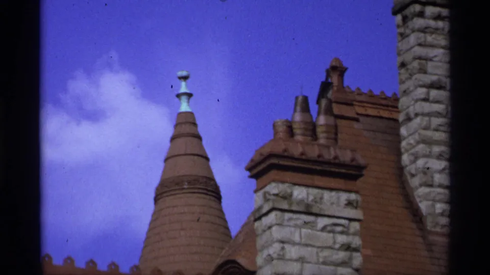 1974 Vancouver home-movie frame focused on roof details, including a conical turret, stone chimney, decorative ridge, and blue sky.
