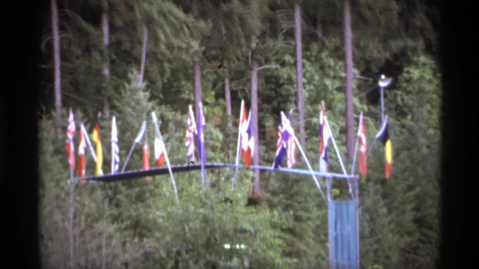 Soft 1974 Vancouver 8mm frame of flags in a wooded outdoor display, with dense greenery behind them.